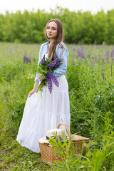 Beautiful young girl in a white dress, straw hat with a bouquet of violet flowers in her hands and picnic basket. Pretty woman in summer in the blooming field holding a bunch of purple lupin