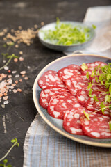 Slices of smoked cervelat salami sausage with spinach microgreen on black, side view, selective focus.