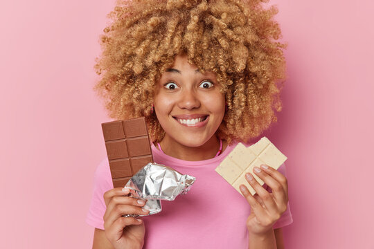 Positive curly haired woman holds two bars of sweet chocolate has sugar addiction bites lips looks with happy surprised expression isolated over pink background. Female suggests appetizing snack