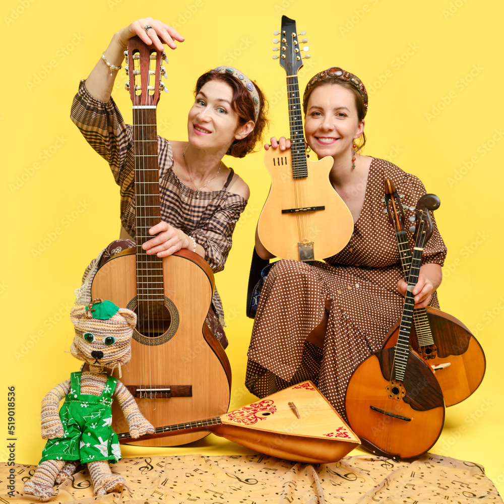 Wall mural Women musicians in dresses with musical instruments on a yellow studio background. Happy artists with stringed musical instruments with smiles on faces