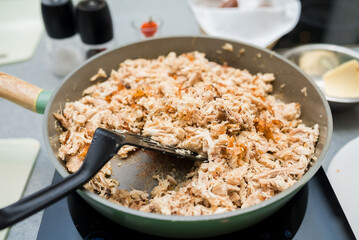 chicken meat fibers are fried in a pan on an electric stove, stirred with a black plastic spatula