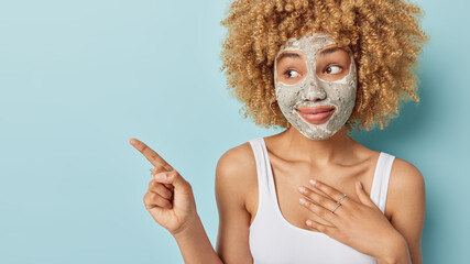 Studio shot of good looking woman with curly hair applies nourishing clay mask to enhance elasticity keeps hand on chest dressed in casual white t shirt isolated on blue background. Beauty procedures