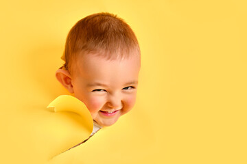The cunning face of a child looks out of a hole in the studio yellow background. An insidious baby...
