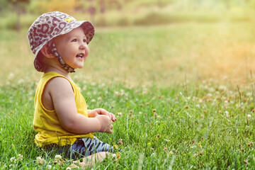 Happy toddler baby boy sit on green grass in nature. Smiling child in shorts, yellow t-shirt and hat plays in the summer in the park. Kid age one year