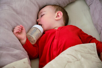 Toddler baby sleeps in a crib with a bottle of milk formula in his mouth. Child boy in red clothes sleeping with food