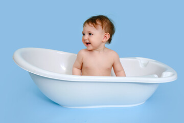 Naklejka premium Happy baby toddler boy is sitting in a white tub on a studio blue background. A smiling child at the age of one year, copy space