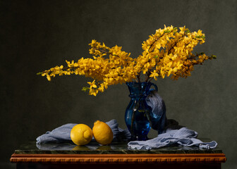 A bouquet of yellow flowers in a blue vase on a marble table. A blue handkerchief and two lemons. Flowering branches of forsythnia Wooden table on a gray background. Spring mood and aroma.