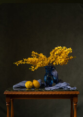 A bouquet of yellow flowers in a blue vase on a marble table. A blue handkerchief and two lemons. Flowering branches of forsythnia Wooden table on a gray background. Spring mood and aroma.2
