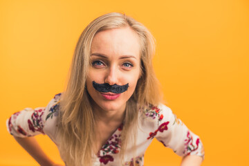 Closeup portrait of a young hilarious European girl with artificial facial hair. Isolated copy space over orange background. Woman with fake moustache bending over to the camera. High quality photo