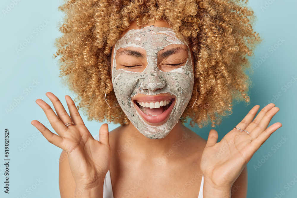 Canvas Prints Positive curly haired young woman laughs gladfully smiles broadly keeps palms raised applies facial beauty mask for skin treatment expresses happy emotions poses indoor against blue background