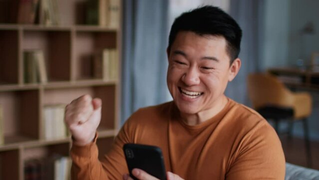 Great news concept. Emotional middle aged asian man web surfing on smartphone and enjoying win, laughing at home