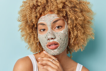 Close up portrait of curly woman touches jawline applies nourishing clay mask to reduce blackheads looks directly at camera takes care of facial skin isolated over blue background. Facial treatments