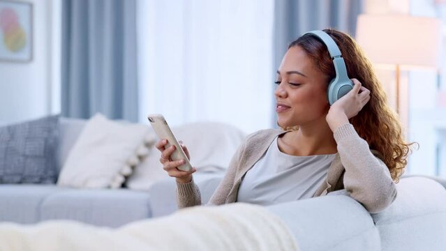 Woman listening to music on her phone using headphones, nodding her head with increased serotonin in home living room. Relaxed woman browsing song playlist on technology and relaxing on sofa