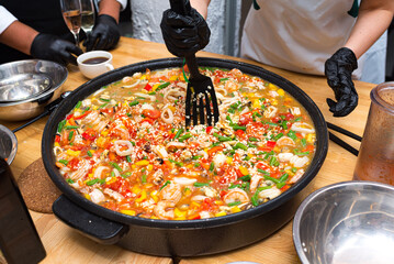 A girl is stirring paella with a black plastic spatula. A large paella bowl on the table. A paella of seafood cocktail, green string beans, red and yellow bell peppers, and broth.