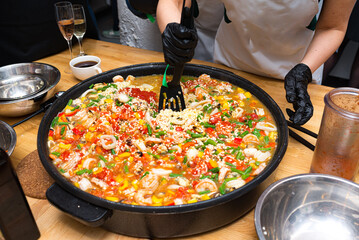 A girl is stirring paella with a black plastic spatula. A large paella bowl on the table. A paella of seafood cocktail, green string beans, red and yellow bell peppers, and broth.