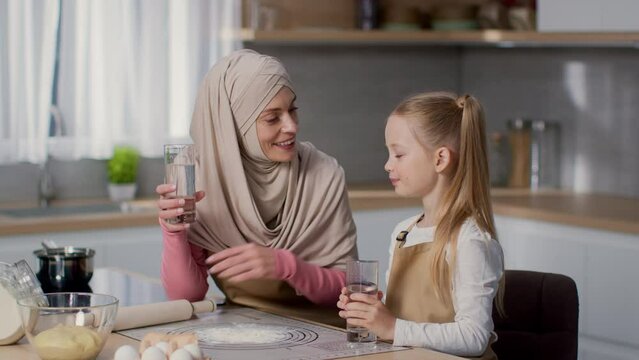 Healthy habits in everyday life. Happy muslim mother and her cute little daughter drinking water at kitchen, free space