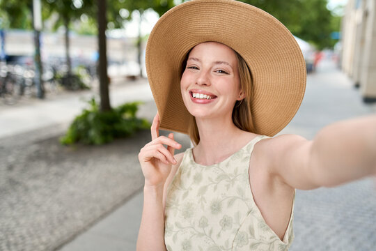 Young happy woman wearing hat taking selfie on summer city street. Cheerful pretty lady fashion model holding smartphone looking at mobile camera making portrait photo outdoor.