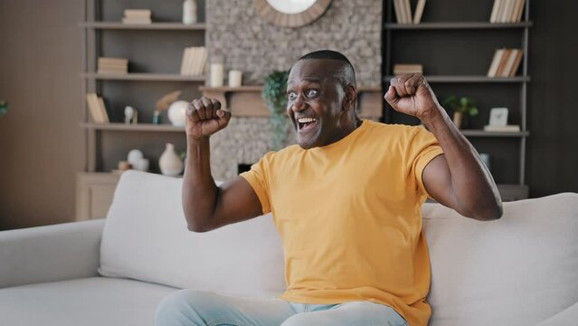 Senior football fan african american man bachelor sit on cozy sofa in apartment watch TV sport program hockey championship rooting for favorite team on television rejoice victory triumph yes gesture
