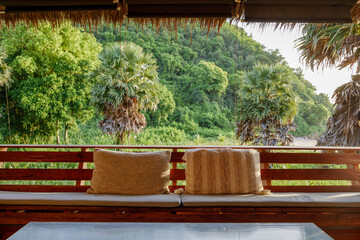 Cozy and comfortable wooden sofa and textile pillows with a scenery of tropical forest atmosphere and sunshine. Summer asian tropical resort vibe.	