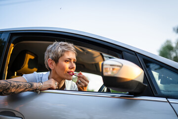 One woman mature caucasian female businesswoman sitting in car putting lipstick fixing repairing makeup on her face while waiting in summer day evening real people copy space gray hair