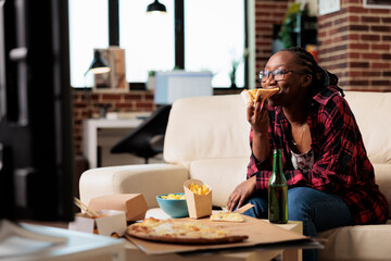 African american woman eating slice of pizza and watching movie, relaxing on couch with takeaway food delivery. Enjoying fast food meal from takeout package to watch film on television.
