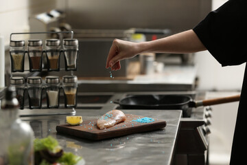 Female chef adding blue salt to raw chicken fillet in restaurant kitchen, closeup. Cooking food
