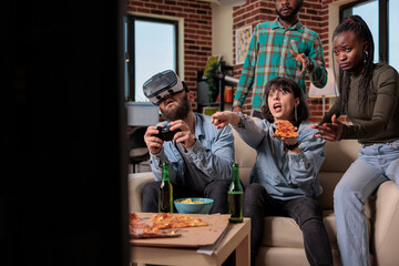 Multiethnic group of people having fun with vr glasses and video games, playing competition with augmented reality goggles. Friends feeling happy together at party gathering with alcohol.