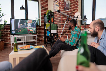 Diverse group of people playing shooting video games on tv console, drinking beer at social gathering with friends. Having fun with gaming competition and leisure activity at house party.