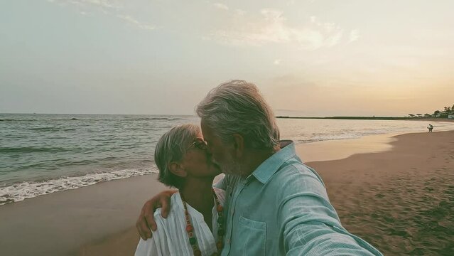 Close up of two cute and happy seniors having fun and enjoying together a sunset day at the beach. Mature couple in love kissing together with the sunset at the background.
