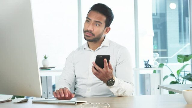 Young business man reading a message on his phone and working on his computer in the office. Handsome corporate professional sending an email and updating the project deadline after receiving a text