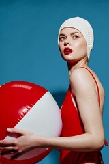 Delighted woman in a bathing cap red swimsuit with a striped ball turns back posing on a blue...