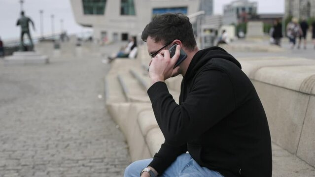 Young mid-twenties white male using mobile phone gets up and walks away in cinematic slow motion. Mid-wide shot showing Liverpool in background.