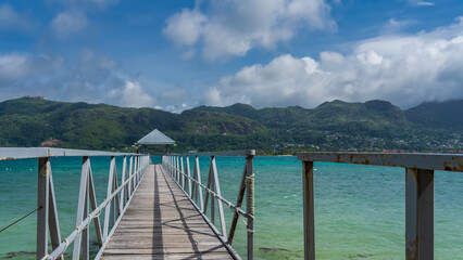 A wooden pedestrian walkway with metal railings runs over the turquoise ocean. There is a canopy in front. Green mountains of the island against a blue sky with clouds. Seychelles