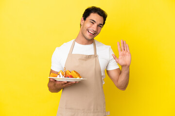 Restaurant waiter holding waffles over isolated yellow background saluting with hand with happy...