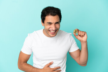 Young man holding a Bitcoin over isolated blue background smiling a lot