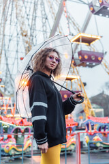 Obraz na płótnie Canvas Portrait of a young stylish woman with curly hair and colored make-up posing with transparent umbrella in amusement park. Street fashion