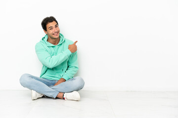 Caucasian handsome man sitting on the floor pointing back