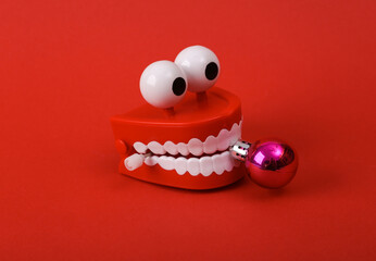 Funny toy clockwork jumping teeth with eyes holding a christmas ball on red background.