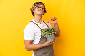 Gardener blonde man holding a plant isolated on yellow background proud and self-satisfied