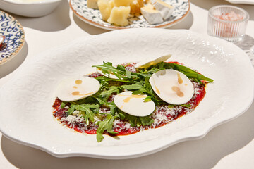 Italian antipasti beef carpaccio with parmesan, ruccola and mushrooms on white table. Carpaccio with raw meat and cheese in italian style with sunny shadows. Italian menu. Aesthetic food composition.