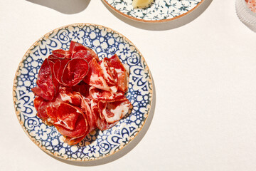 Meat antipasti on ceramic plate on white table with hard shadow. Meat platter with bresaola, hamon, prosciutto. Meat appetizer in Italian menu. Aesthetic food composition. Sunny day in tuscany.