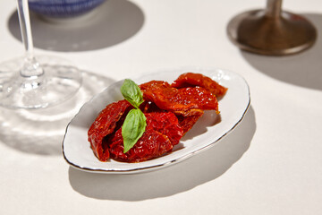 Italian antipasti - sundried tomatoes in ceramic dish on white table. Dried tomato in olive oil with basil on summer day. Appetizer from dried tomatoes in italian restaurant menu.