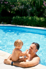 Smiling dad stands in the water and hugs a small child sitting on the edge of the pool