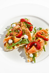 Bruschetta with salmon, avocado and paprika on white plate. Salmon bruschetta on crispy focaccia. Appetizer with guacamole, trout, cheese and paprika on toast. Fish antipasti in restaurant menu.