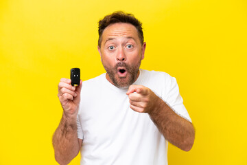 Middle age caucasian man holding car keys isolated on yellow background surprised and pointing front