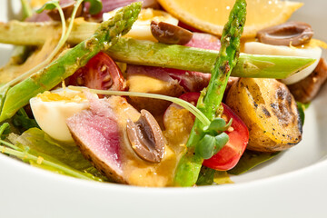 Classic Nicoise salad with grilled tuna, potatoes, asparagus, tomatoes and olives with mustard dressing. French cuisine - Tuna salad with vegetables, eggs and vinaigrette sauce in white background.