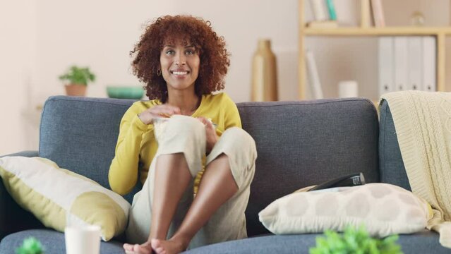 Woman watching her favorite TV show while enjoying popcorn at home. Young modern girl relaxing on the sofa and watching a funny film. A fun weekend staying indoors and snacking alone on the couch