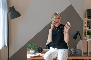 Smiling business woman using phone in office. Small business entrepreneur looks up talking on a cell phone and smiles. Lounging on the desk in a black T-shirt and white trousers