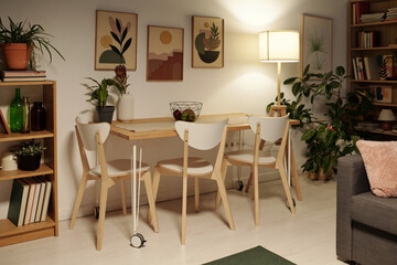 Several wooden chairs standing around dinner table by wall with group of paintings and shelves with books in spacious living room