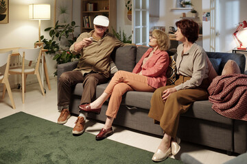 Senior man with vr headset sitting on couch next to two aged women in living room and telling them what he sees in extended reality
